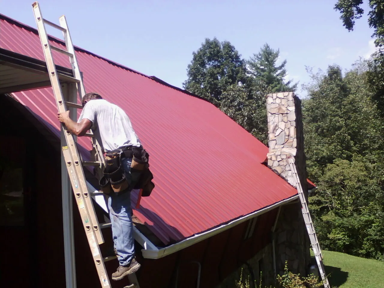 Roofer on a ladder beside a red metal roof and stone chimney — Soffit & Fascia Repair by Roofing Brothers in Paxico